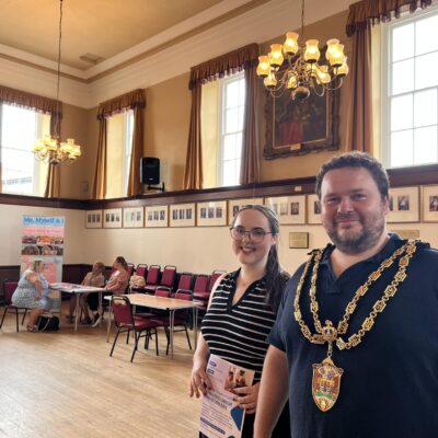 Neath Town Mayor wearing his chains and Carys Hope - Community Engagement Officer - standing in Neath Town Hall. Tables and chairs behind, with people sitting